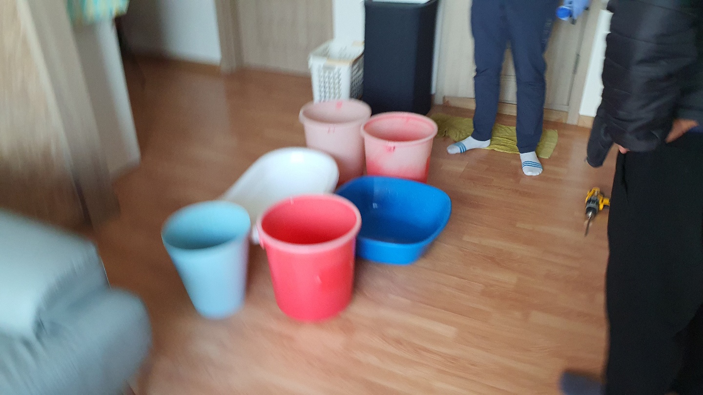 거실 나무 바닥 위에 다양한 색상의 대야와 양동이가 놓여 있고 전동 드릴과 함께 보수 공사가 진행 중인 모습입니다.