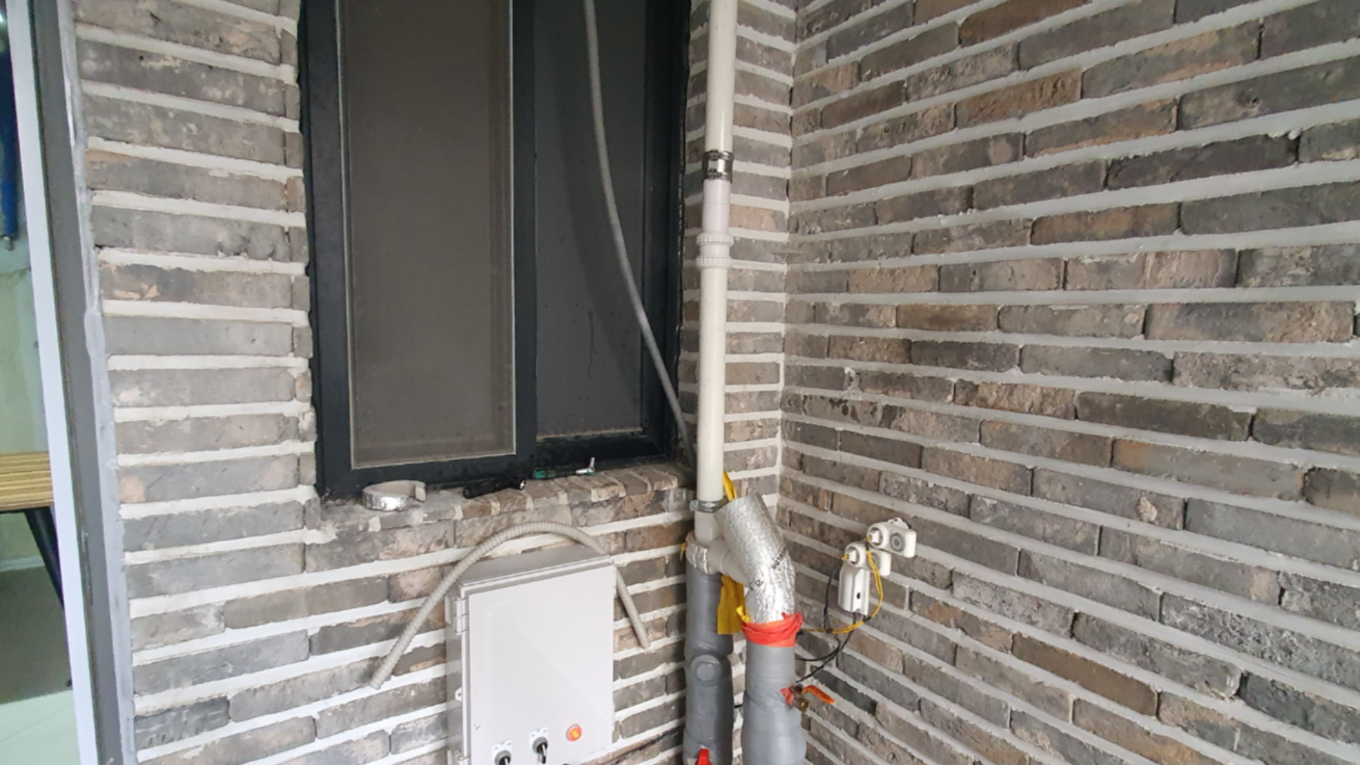 벽돌 벽면에 설치된 흰색 PVC 우수관 하단부에 임시방편으로 테이핑 처리가 되어 있으며 옆에 전기 분전함이 보입니다.